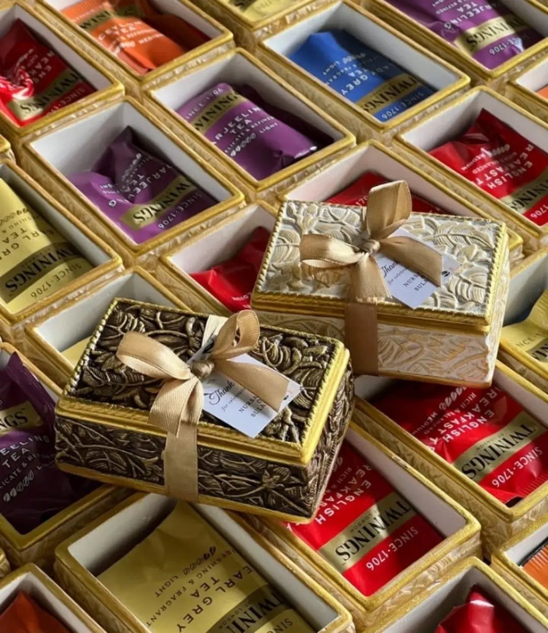 Tea Bag Gift Ideas to Make or Buy: 11 Sweet Picks 3 Ornate gift boxes resting on gold-rimmed dishes filled with colorful tea sachets.