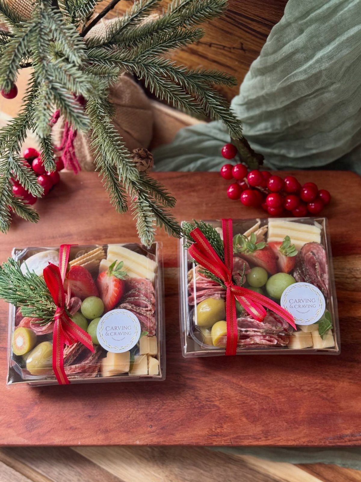 12 Thanksgiving Gifts for Coworkers They'll Actually Use 10 Charcuterie gift boxes with red ribbons on a wooden board with evergreen branches.