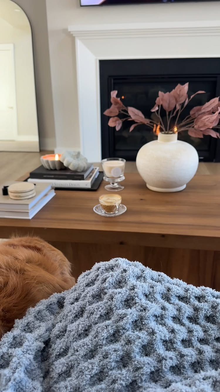 11 Thanksgiving Gifts for Family, From Candles to Cozy Throws 11 Grey knit blanket and candles on a wooden table in front of a fireplace