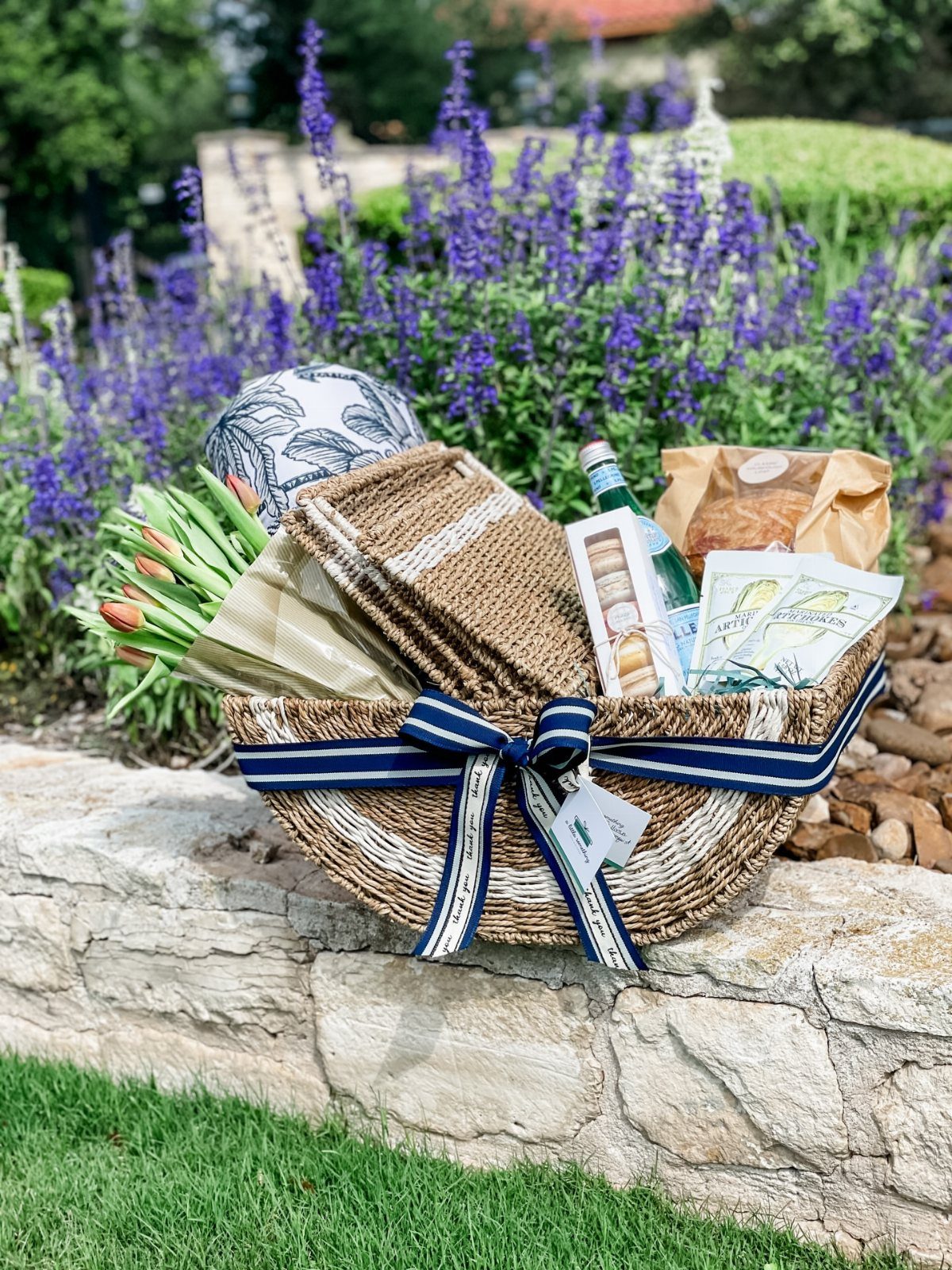 Woven gift basket with blue ribbon, snacks, and tulips on a stone wall.