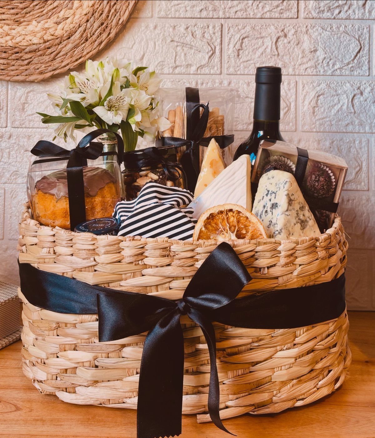 Woven gift basket with red wine, assorted cheeses, crackers, and white flowers.
