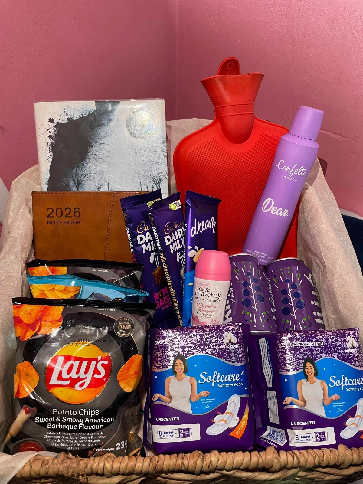 Wicker gift basket containing a red hot water bottle, snacks, and personal care items.