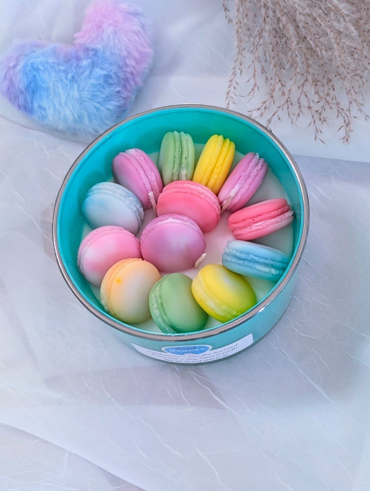 Colorful macaron-shaped candles arranged inside a turquoise round tin on white fabric.