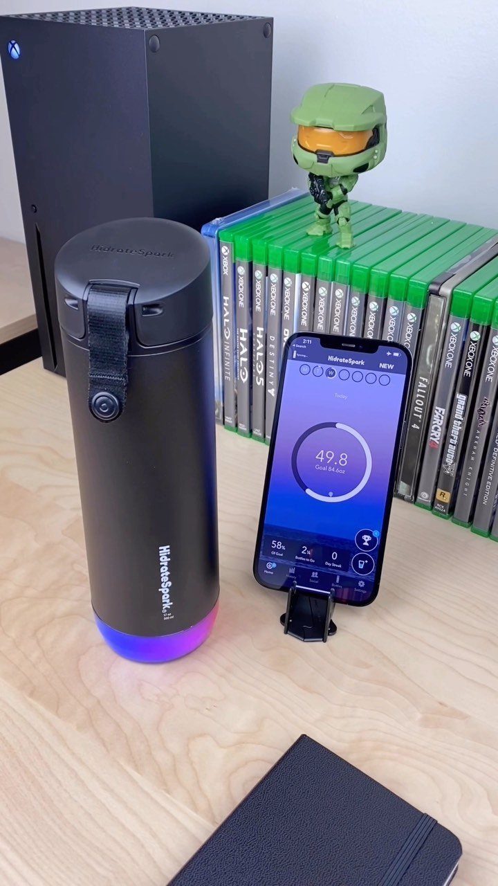 Smart water bottle with glowing base next to a smartphone and gaming gear.