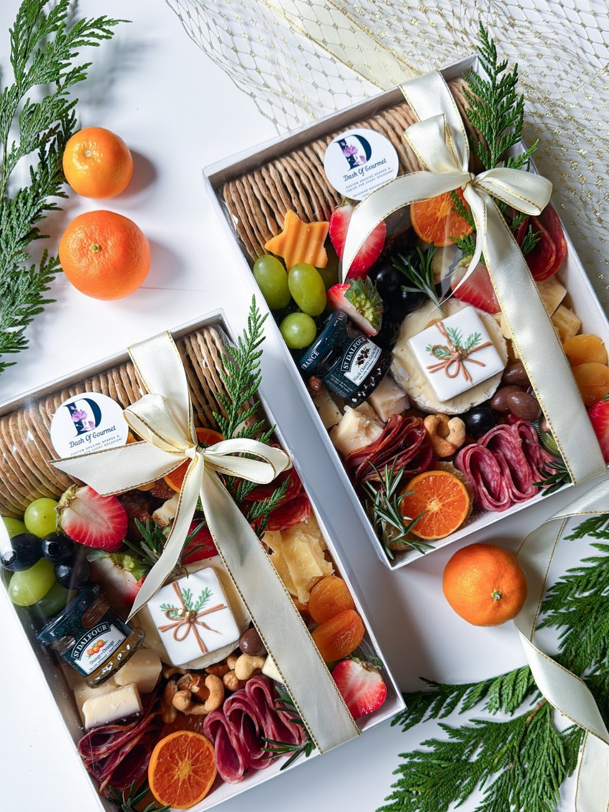 Two charcuterie gift boxes with meats, cheeses, and fruits tied with cream ribbons.