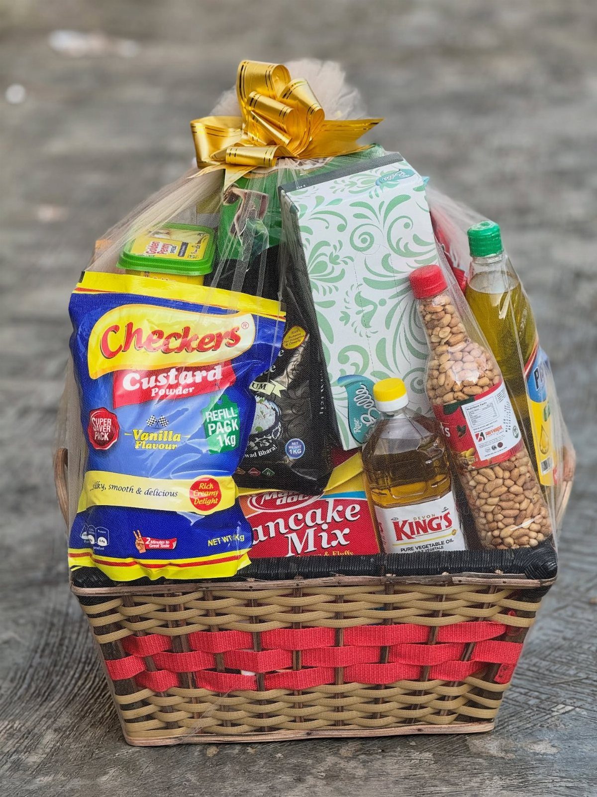 Woven gift basket filled with food items wrapped in cellophane with a gold bow.