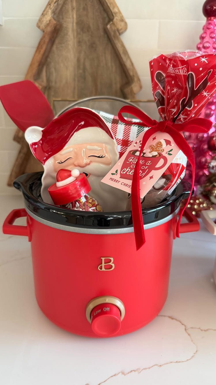 Red slow cooker filled with Christmas treats, a Santa figurine, and a red ribbon.